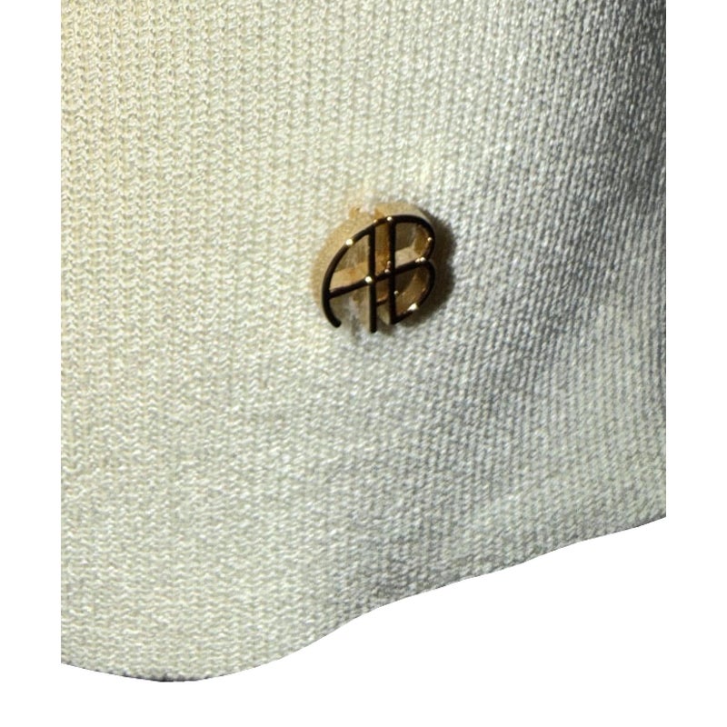 Close-up of the gold-tone AB emblem and ivory knit fabric near the ANINE BING sweater hem.
